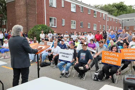 A speaker at a podium addressing seated co-op residents holding coalition signs